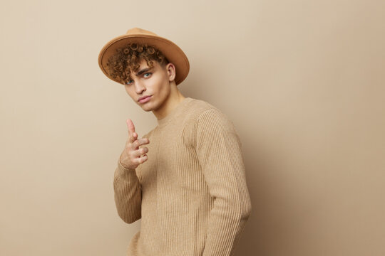 A Charming Man In A Beige Sweater Stands On A Beige Background With A Hat On His Head And Looks Pleasantly Into The Camera Holding Out His Hand