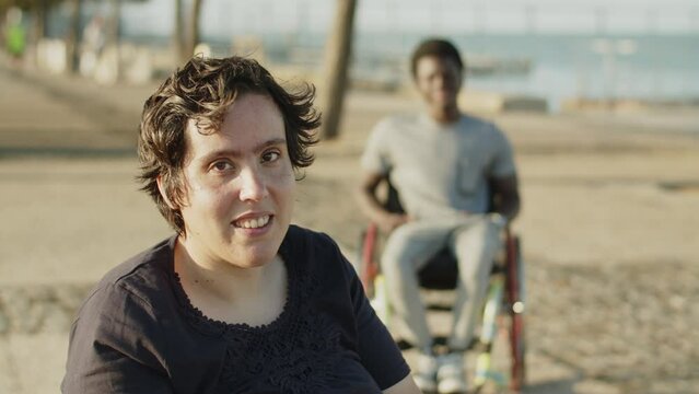 Portrait of young woman with disability smiling at camera outdoors with male friend using wheelchair in background. Happy woman walking in park. Woman with disability concept