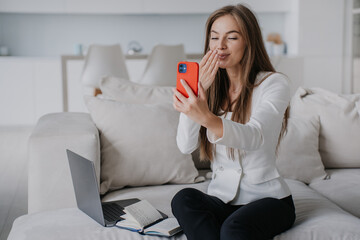 Gorgeous businesswoman in white jacket and black pants makes video call using phone sends air kiss to husband sits  on cozy couch with laptop at home. Cheerful young pretty blonde woman remote working