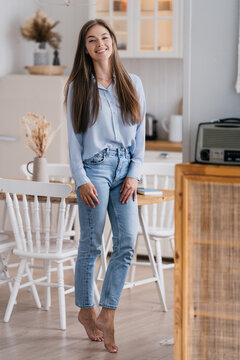 Vertical Shoot Of Gorgeous Blonde Young Woman In Casual Posing At Kitchen Smiling Wide Looks At Camera Happily Stands At Kitchen. Beautiful Caucasian Fit Girl  With Long Loose Hair At Home.