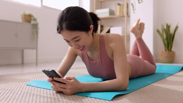 Smiling young asian woman using mobile phone while relaxing after exercising at home. Female fitness girl ying on mat checking smartphone. Sport, technology and healthy lifestyle concept.