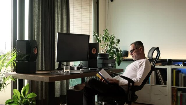 Handsome middle age man reading book at home by his computer desk near the window