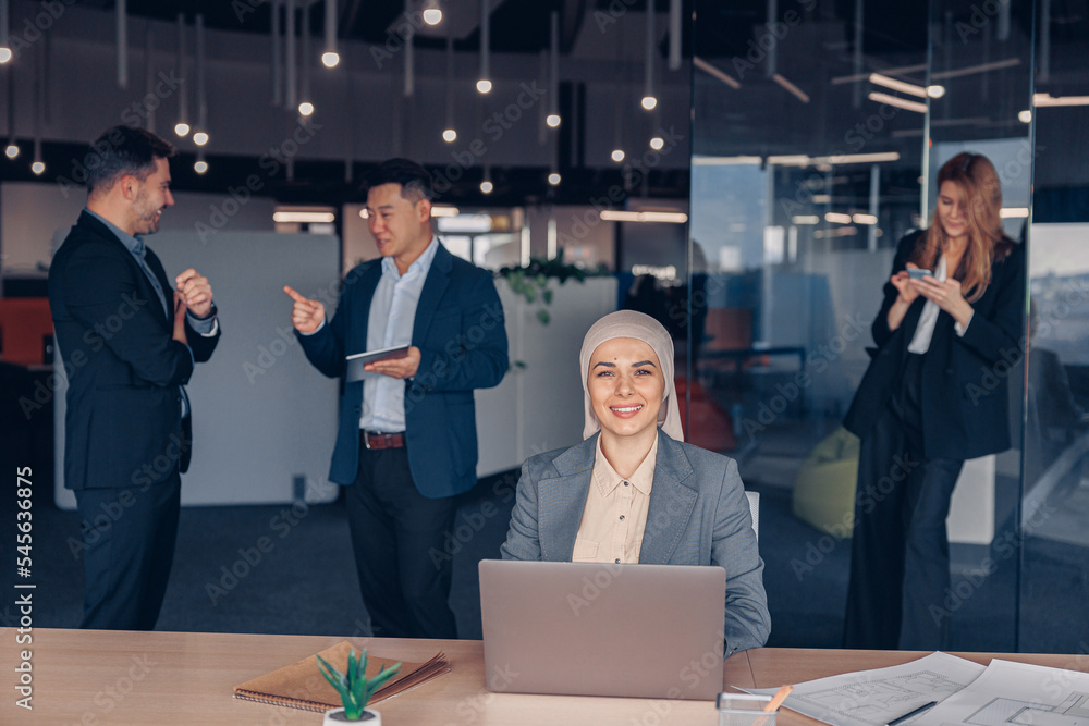 Wall mural smiling muslim businesswoman in hijab working on computer while sitting in modern office