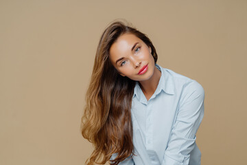 Obraz premium Portrait of pleasant looking female tilts head on right, has long well cared hair, wears blue shirt, has appealing appearance, makeup, isolated over brown background with blank space for information