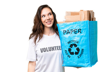 Young caucasian woman holding a recycling bag full of paper to recycle over isolated background looking up while smiling