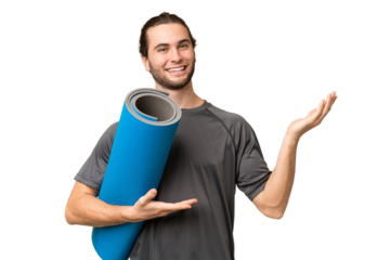 Young sport man going to yoga classes while holding a mat over isolated background extending hands to the side for inviting to come