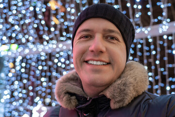 Photo of attractive millennial man located next to Christmas decorations lights on street at winter. Happy man in winter clothes at christmas market. Festive garland lights.