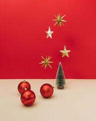 Golden star shaped ornament flying above small Christmas tree decorated with big red baubles on red and beige background. Aesthetic vibrant New Year or Christmas holiday concept.