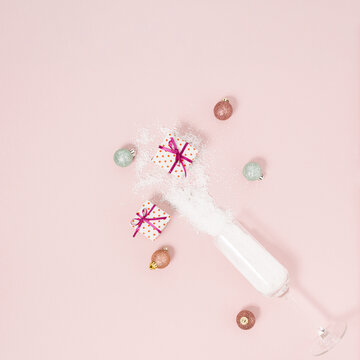 Minimal composition with champagne glass, snow, pink and green baubles, wrapped present against pastel pink background. Creative Christmas decoration with gift box. New Year party aesthetic style.