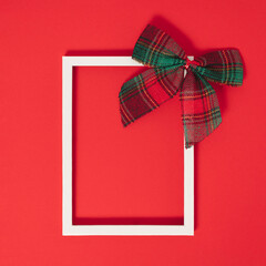 White frame with plaid bow on top on red background. Minimal Christmas greeting card. 
