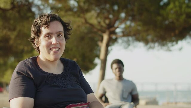 Portrait of young woman with disability smiling at camera in park with male friend using wheelchair in background. Happy woman walking in park. Woman with disability concept