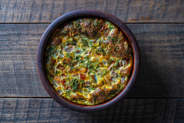 Fototapeta na wymiar Ceramic bowl with vegetable frittata, simple vegetarian food, top view. Frittata with egg, tomato, pepper, onion, parsley and cheese on wooden table. Italian egg omelette