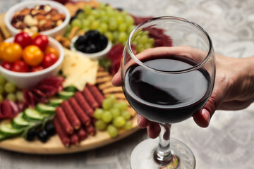 A person's hand elegantly holds a glass of red wine, with a background of assorted cheeses and charcuterie on a wooden board. The epitome of a fine dining experience