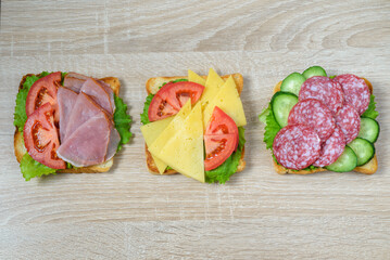 Sandwiches with ham, salami, cheese, tomato and cucumber on wooden desk.
