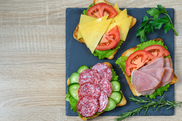 Sandwiches with ham, salami, cheese, tomato and cucumber on slate board.