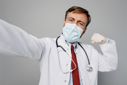 Close up male doctor man wears white medical gown suit mask work in hospital do selfie shot pov on mobile cell phone show muscles isolated on plain grey background studio. Healthcare medicine concept.