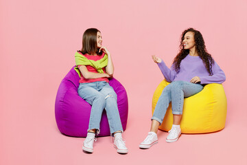 Naklejka premium Full body young two friends positive happy fun cheerful women 20s wears green purple shirts together sit in bag chair talk speak isolated on pastel plain light pink color background studio portrait.