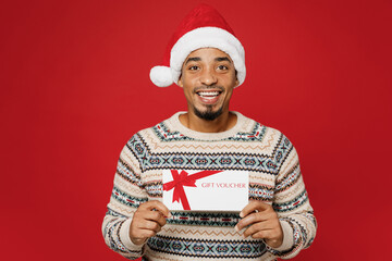 Merry young man wear warm cozy Christmas sweater Santa hat posing hold gift certificate coupon voucher card for store isolated on plain red background. Happy New Year 2023 celebration holiday concept.