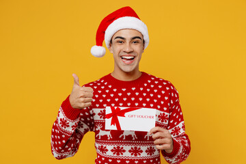 Merry young man wear red knitted christmas sweater Santa hat posing hold gift certificate coupon voucher card for store show thumb up isolated on plain yellow background. Happy New Year 2023 concept.