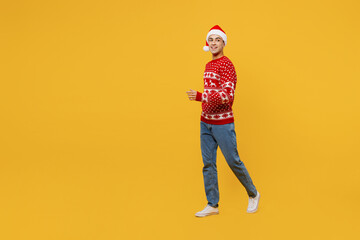 Fototapeta premium Full body side view merry young man wear red knitted Christmas sweater Santa hat posing walk go look aside on area isolated on plain yellow background. Happy New Year 2023 celebration holiday concept.