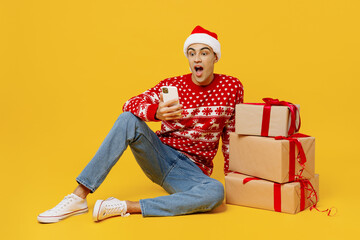 Full body merry young man wear red Christmas sweater Santa hat posing sit near red present boxes gift ribbon bow use mobile cell phone isolated on plain yellow background. Happy New Year 2023 concept.