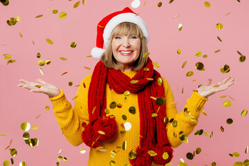 Merry elderly woman 50s years old wear yellow sweater red scarf Santa hat posing spread hands catch confetti glitters rainfall isolated on plain pink background. Happy New Year Christmas 2023 concept.