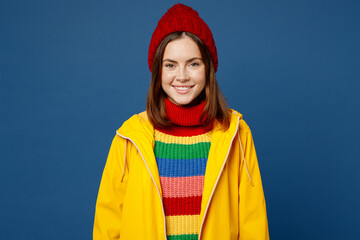 Young smiling happy cheerful fun woman wears sweater yellow waterproof raincoat outerwear red hat look camera isolated on plain dark royal navy blue background Outdoor wet fall weather season concept