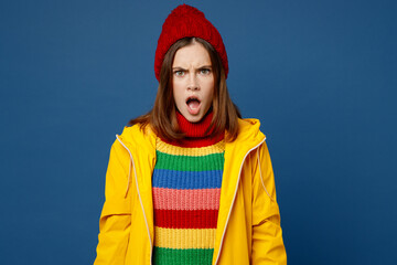 Young woman in sweater red hat yellow waterproof raincoat outerwear look camera with open mouth scream shout isolated on plain dark royal navy blue background Outdoor wet fall weather season concept.