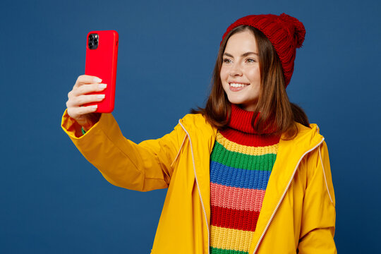 Young woman in sweater red hat yellow waterproof raincoat outerwear do selfie shot on mobile cell phone post isolated on plain dark royal navy blue background Outdoor wet fall weather season concept