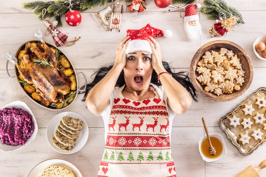 Stressed female cook in a Christmas apron and Santa hat holding her head and lying on the floor surrounded by gingerbread, Linz cakes, on the other side: roast goose or turkey with side dishes