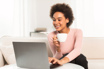 Happy millennial african american woman enjoy cup of coffee and typing on laptop in bright living room