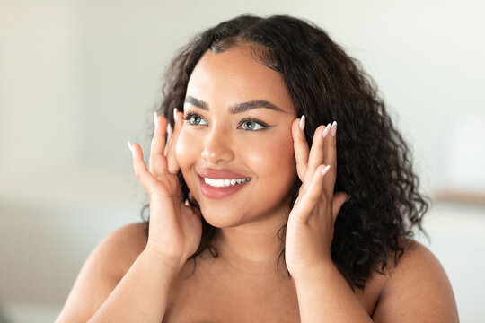 Beautiful black chubby lady touching her face and smiling while making beauty treatments at home, enjoying selfcare
