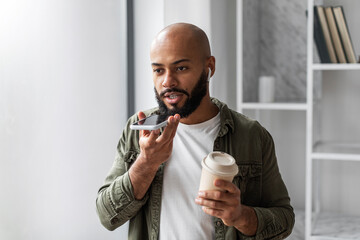 Latin man recording voice message on smartphone and drinking coffee from takeaway cup, enjoying modern technologies