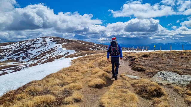 Rear view of man with backpack on hiking trail from Ladinger Spitz to Gertrusk, Saualpe, Lavanttal Alps, Carinthia, Austria, Europe. Trekking on cloudy spring day. Hat in colors of Carinthian flag