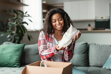 Glad happy black young lady blogger in casual unpacks cardboard box, enjoy fashion sneakers, shows new shoes