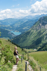 Fototapeta na wymiar Side view of an adult blonde woman with backpack standing on a mountain trail with a lake and the Swiss Alps in the background.