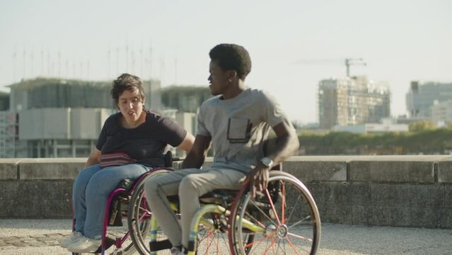 Happy couple using wheelchairs dancing and spinning outdoors. Young African American man and Caucasian woman having fun at quay. Couple with disability concept