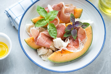 White plate with slices of cantaloupe melon, prosciutto, torn mozzarella and fresh basil, middle close-up on a light-grey granite background