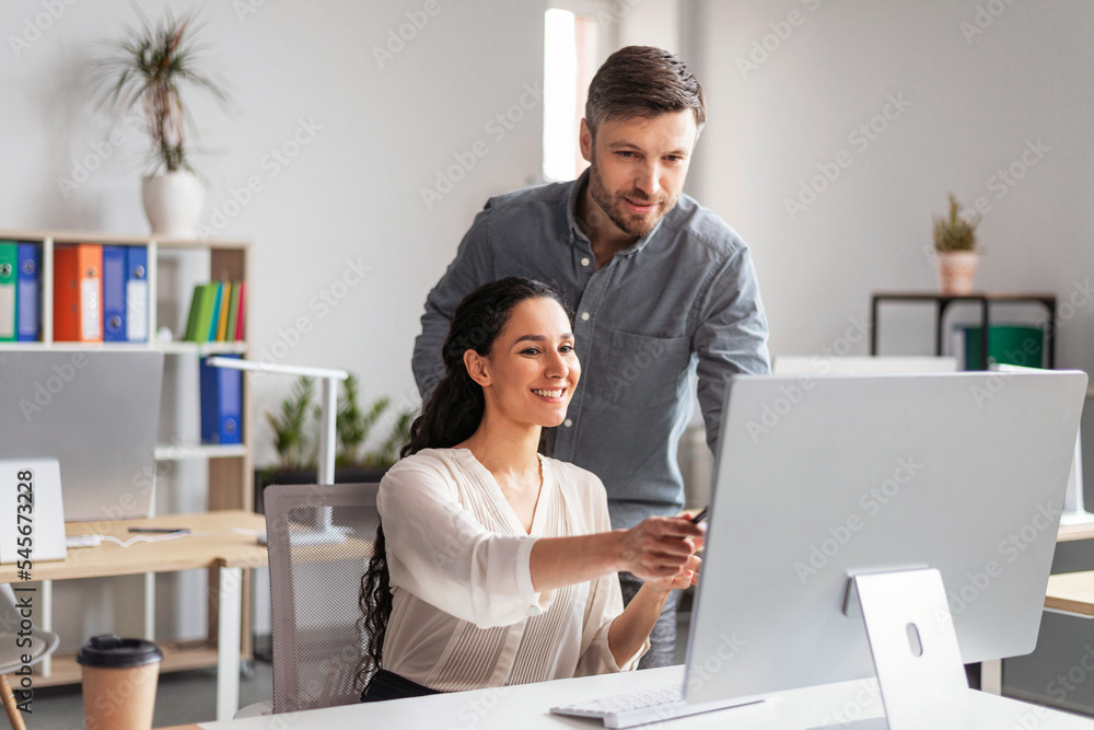 Poster smiling millennial european female showing to man manager with hand in computer screen