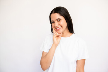 Portrait of happy young woman looking at camera with interest over white background. Caucasian lady wearing white T-shirt touching chin and smiling. Curiosity concept