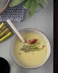 Asparagus cream soup with a spoon inside and some asparagus tips, pink pepper and ham over it on a wooden table.
