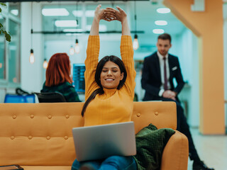 Handsome businesswoman (employee) sitting on a yellow couch while working on her laptop in startup...