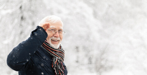 Charismatic elderly gray-haired man in glasses standing with raised hand in a greeting gesture on the street in winter, snow falling, smiling, banner, place for text