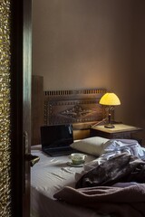 Through a half-open door,  a portable computer and a cup of dirty soup on a plate on the unmade bed of a rustic room lit by a night lamp and the window's sunlight. Interiors full of life.