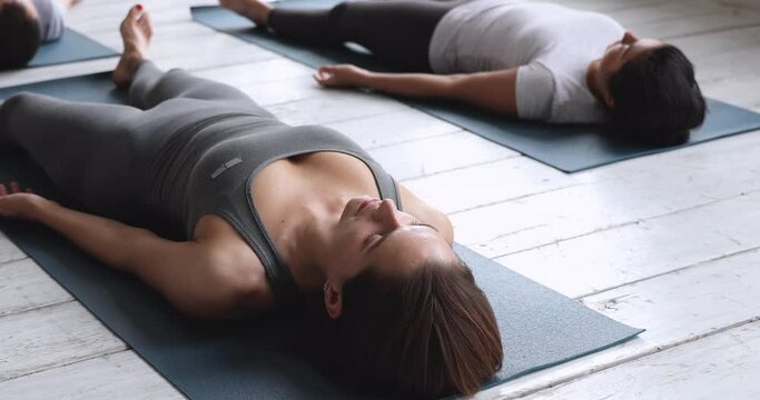 People in sportswear lying down on mats with closed eyes practising Shavasana Dead Body Yoga Posture, close up focus on female session participant or trainer, relax whole body, stress relief concept