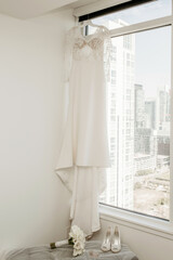 A wedding dress, shoes and a bouquet of the bride are hung by the window in a hotel room against the backdrop of urban development. Wedding accessories of the bride on the wedding day and preparation