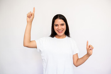 Portrait of happy young woman pointing forefingers upwards over white background. Caucasian lady wearing white T-shirt showing something, looking at camera and smiling. Advertising concept