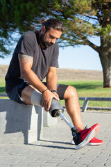 Portrait of man with disability preparing for training. Man sitting outdoors massaging and fixing his prosthetic leg for doing sports. Active life of people with disability, sport activity concept