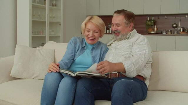 Joyful attractive senior woman and cheerful handsome mature man sitting on couch, watching family photo album, sharing and remembering happy and funny moments while retired couple relaxing at home.