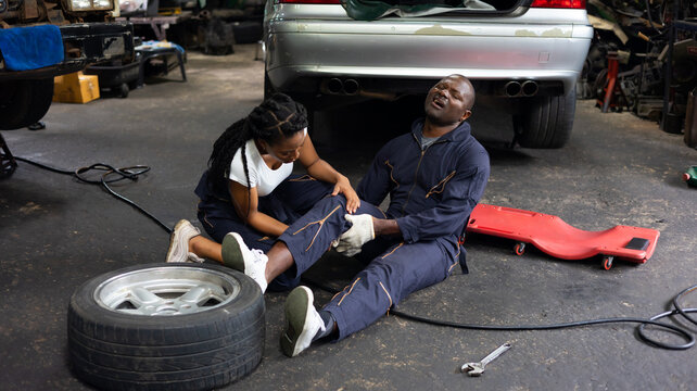 African American woman mechanic helped a black engineer man with knee pain due to work lie on the garage floor.